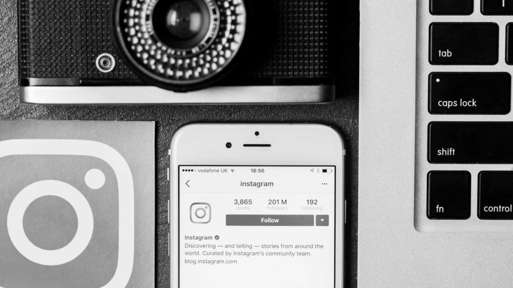 En bas au centre un téléphone sur l'application Instagram. On voit également sur le haut de l'image un appareil photo, en bas sur la gauche le logo d'Instagram et à droite un ordinateur.